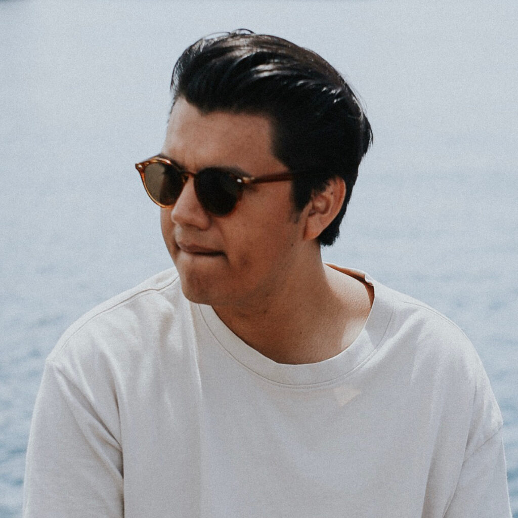 A man with dark hair and a white shirt wearing sunglasses, with the ocean in the background.